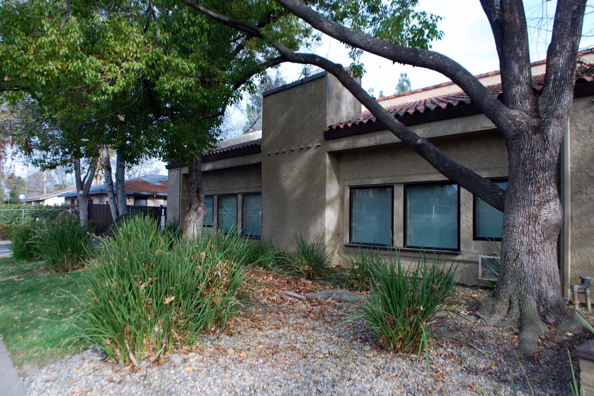 Exterior view of a single-story building with beige stucco walls and a red tile roof, surrounded by trees, bushes, and a gravel landscape area.