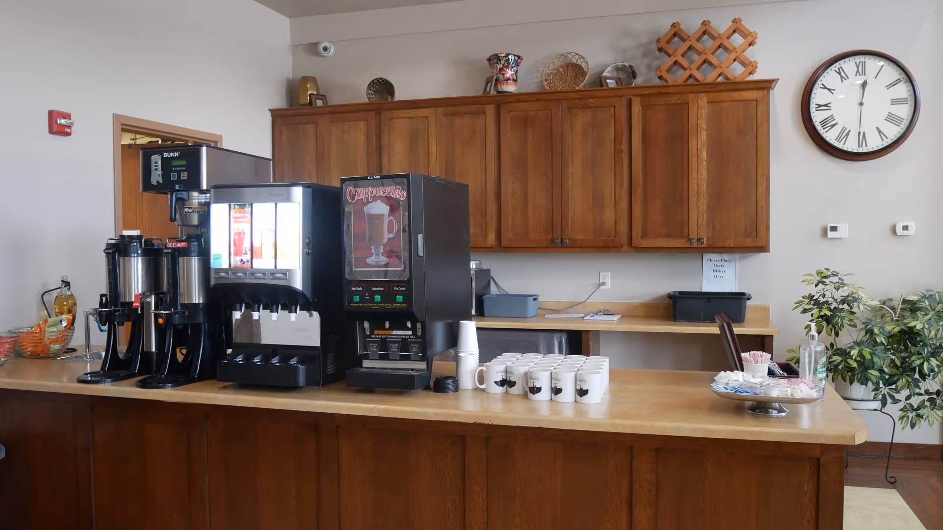 A coffee and beverage station with dispensers, stacked mugs, and wooden cabinets in a common area.
