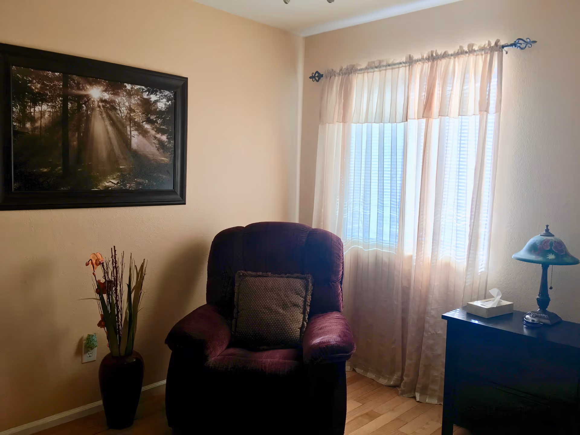 A cozy corner of a room with a dark purple armchair that has a decorative pillow. To the right of the chair is a small black side table with a floral lamp and a tissue box. A window with sheer curtains allows natural light to softly illuminate the space. On the left wall, there is a framed picture of sunlight streaming through trees. A tall vase with artificial flowers and branches is placed on the floor next to the chair.