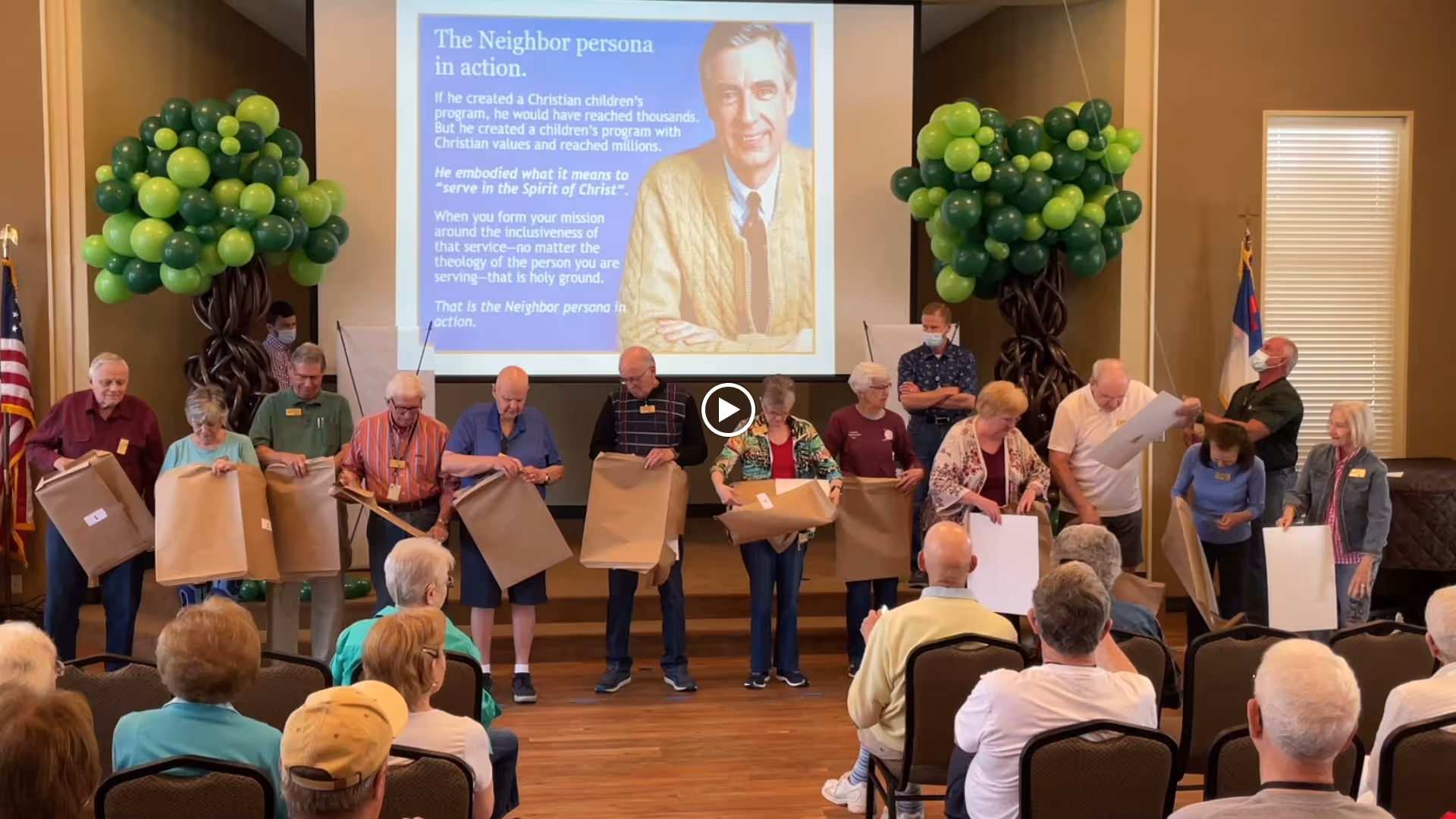 A group of elderly people standing in a row at the front of a room, each holding large sheets of paper or cardboard. Behind them is a large screen displaying an image of Mr. Rogers with text about 'The Neighbor persona in action.' The room has two large balloon decorations shaped like trees on either side of the screen. Several people are seated facing the group, watching the presentation.