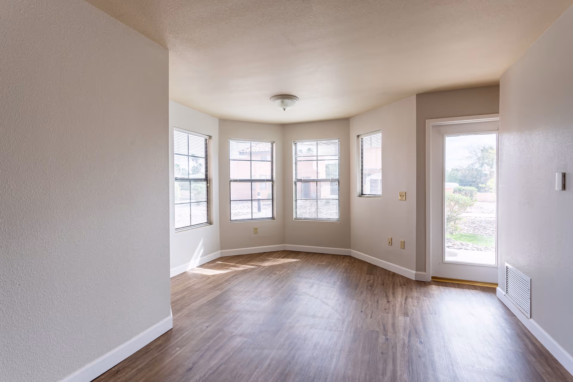 Empty room with light beige walls, wood flooring, multiple windows with blinds, and a glass door leading outside. The room is well-lit with natural light coming through the windows.