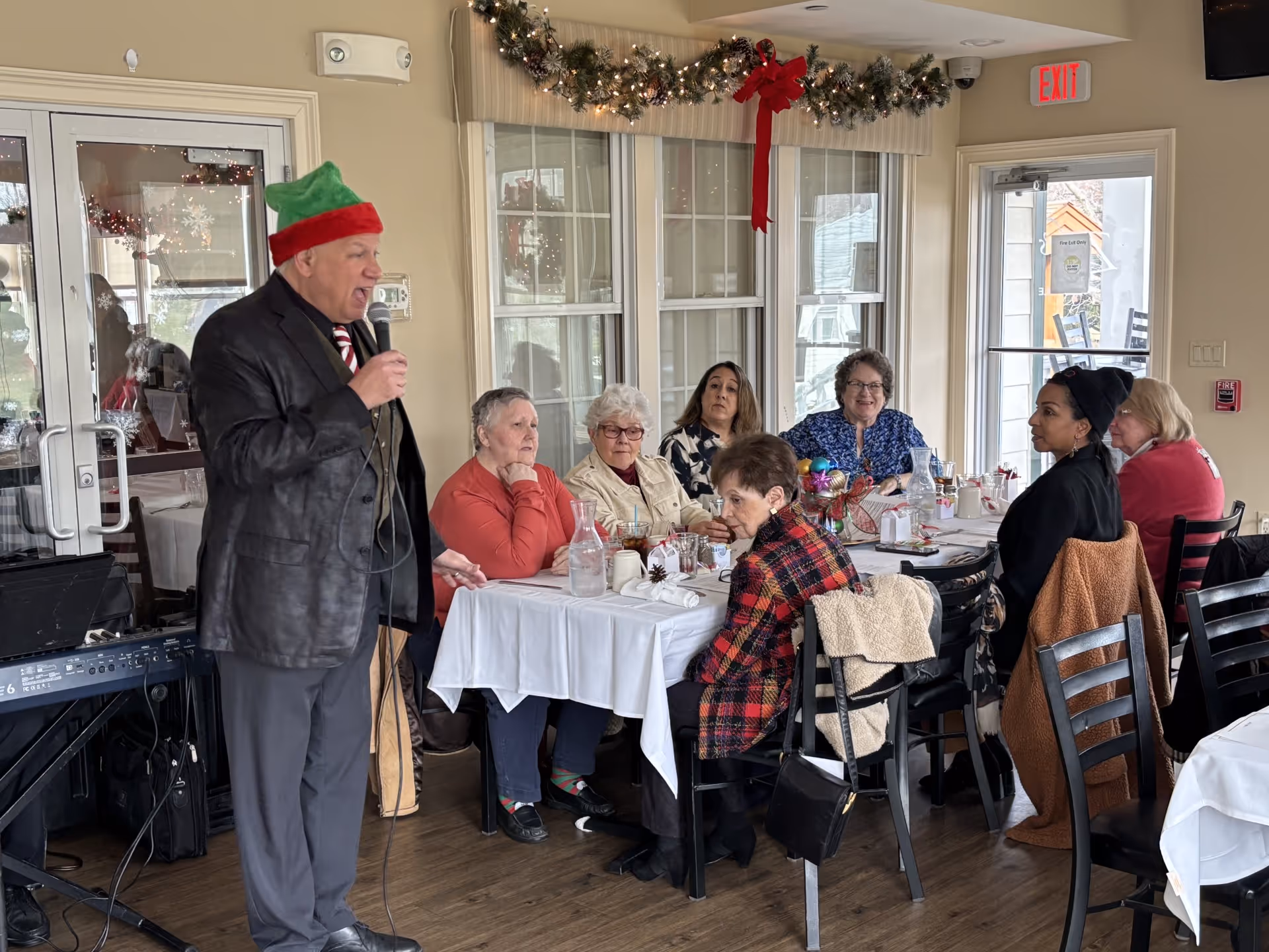 A man wearing a festive hat sings into a microphone while a group of seniors sit around a decorated dining table in a holiday-themed room.