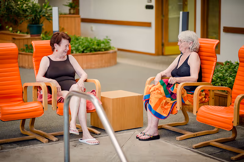 Two elderly women in black swimsuits sitting on orange chairs around a small wooden table near a pool area, one woman has a colorful towel wrapped around her legs, and both are smiling and engaged in conversation.