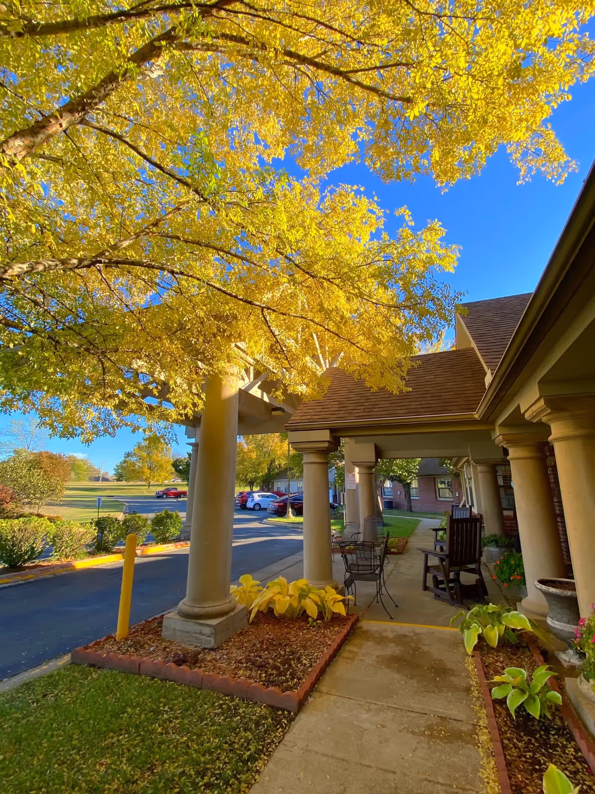 Outdoor view of a senior living facility entrance with large columns supporting a covered walkway. Yellow autumn leaves on a tree overhead, a few chairs and a small table are placed along the walkway. Several parked cars and green bushes are visible in the background under a clear blue sky.