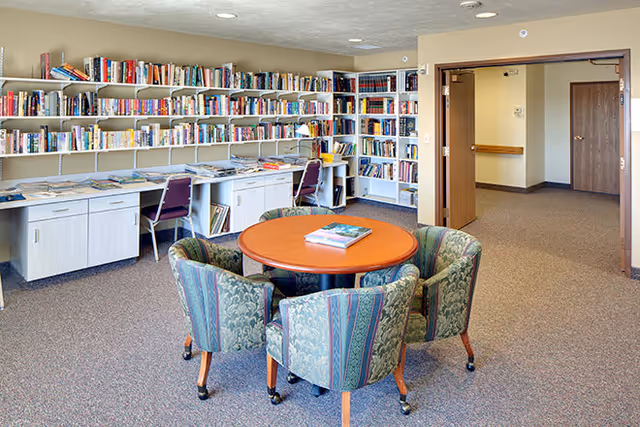 A cozy library room with shelves filled with books along the walls. There is a round wooden table in the center surrounded by four upholstered chairs with patterned fabric. Two desks with chairs are positioned against the wall under the bookshelves. An open doorway leads to a hallway with beige walls and carpeted floors.