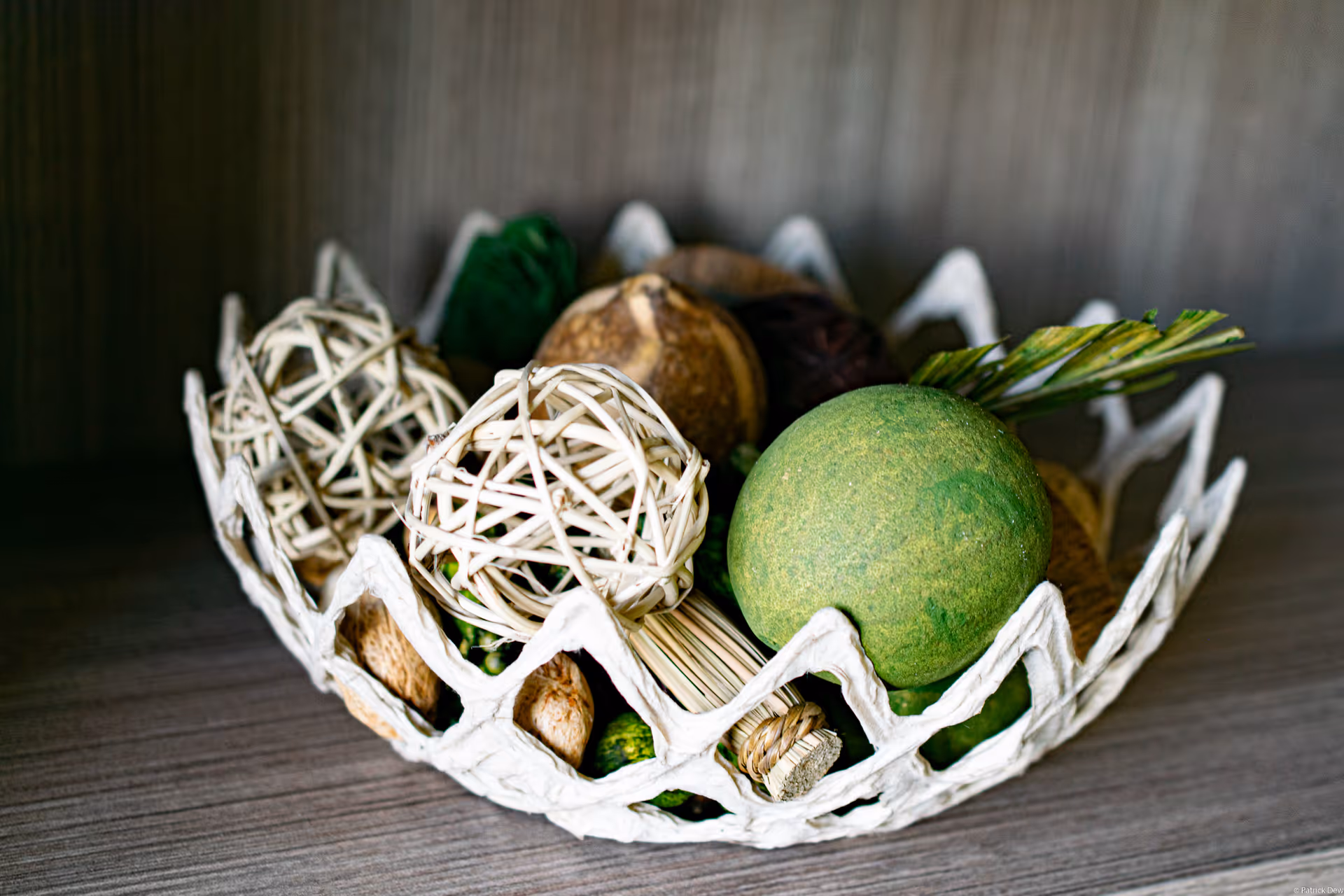 Decorative white woven bowl holding various wicker and green decorative balls on a wooden shelf.
