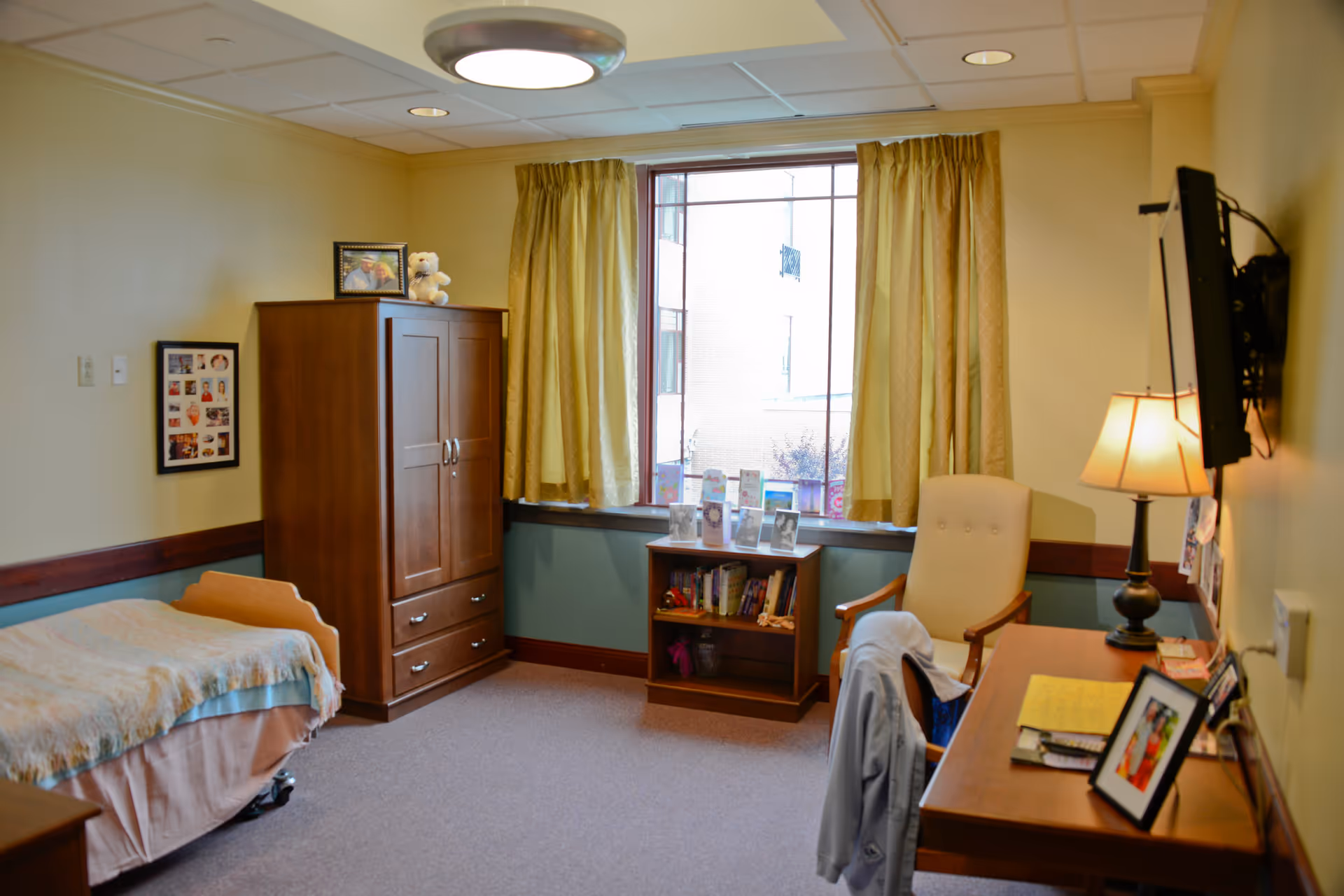 A cozy bedroom in a senior living facility featuring a single bed with a blanket, a wooden wardrobe with a framed photo and teddy bear on top, a window with yellow curtains, a small bookshelf with books and framed pictures, a beige armchair, a wooden desk with a lamp, framed photos, and papers, and a wall-mounted TV.