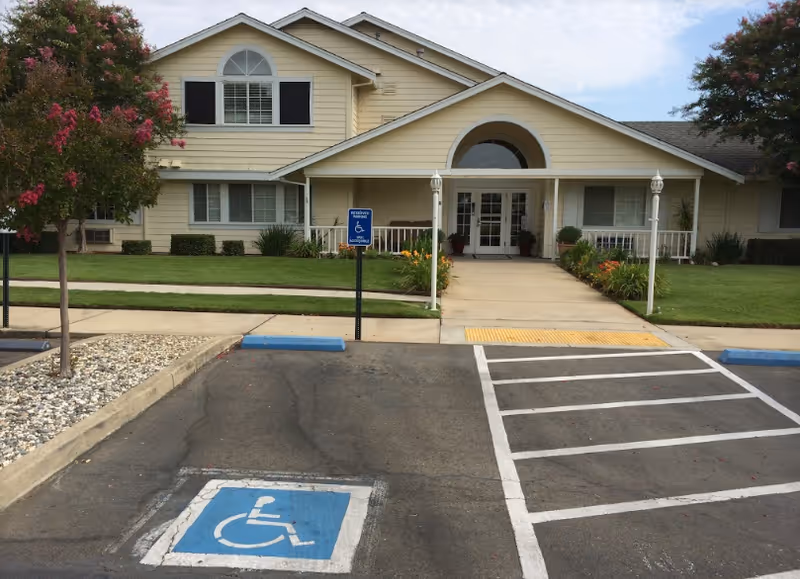 Front exterior view of a single-story assisted living facility building with a beige facade, white trim, and a covered entrance. There is a paved parking area in front with a designated handicapped parking space and signage. The building is surrounded by well-maintained green lawns, flowering trees, and shrubs.