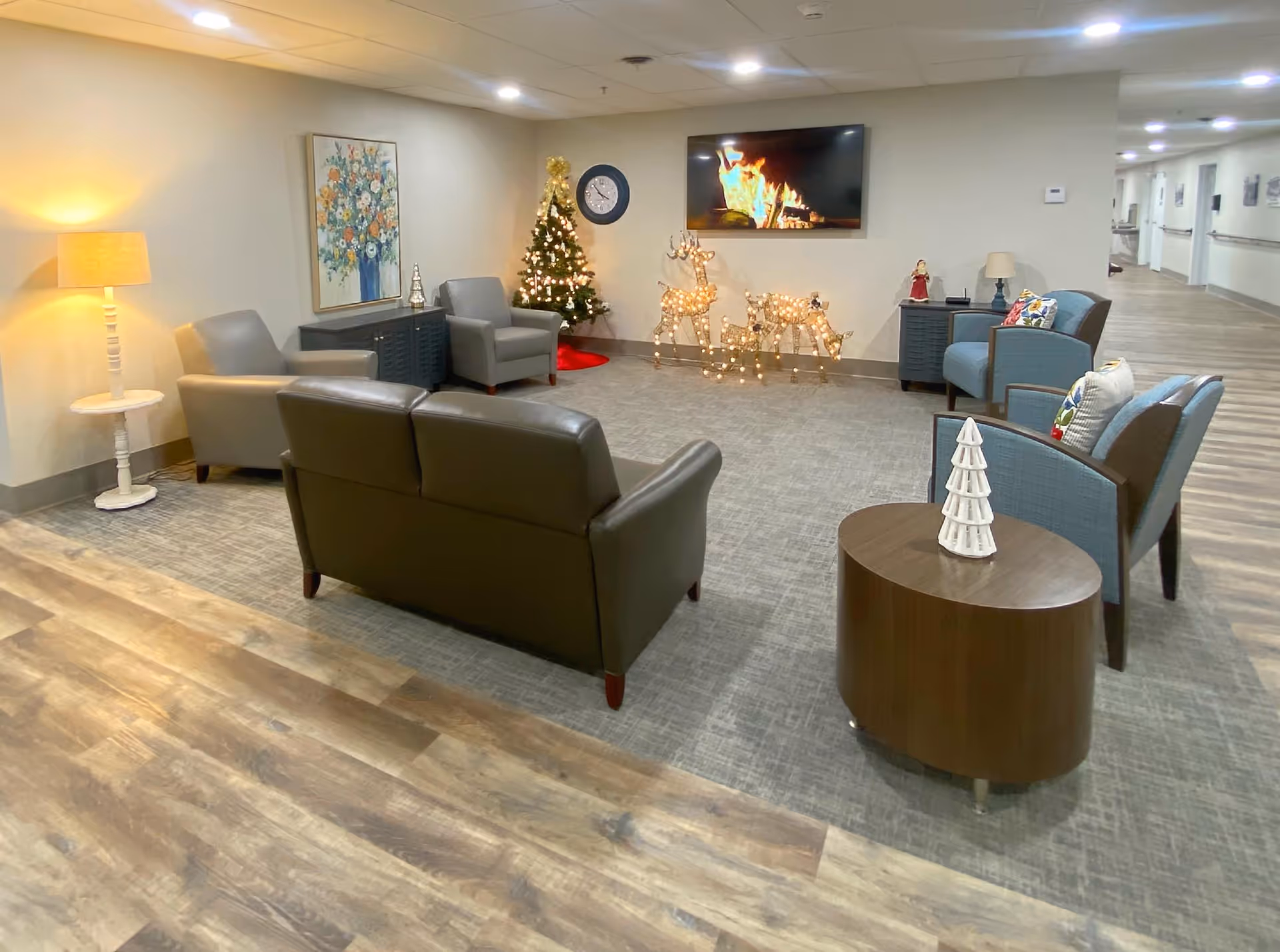 A cozy sitting area in a retirement community decorated for the holidays with a small Christmas tree, lighted reindeer figures, and a wall-mounted TV displaying a fireplace scene. The room features several armchairs, a loveseat, a side table with a small white Christmas tree decoration, a floor lamp, and a floral painting on the wall. The flooring is a combination of wood and carpet, and a hallway extends from the sitting area.