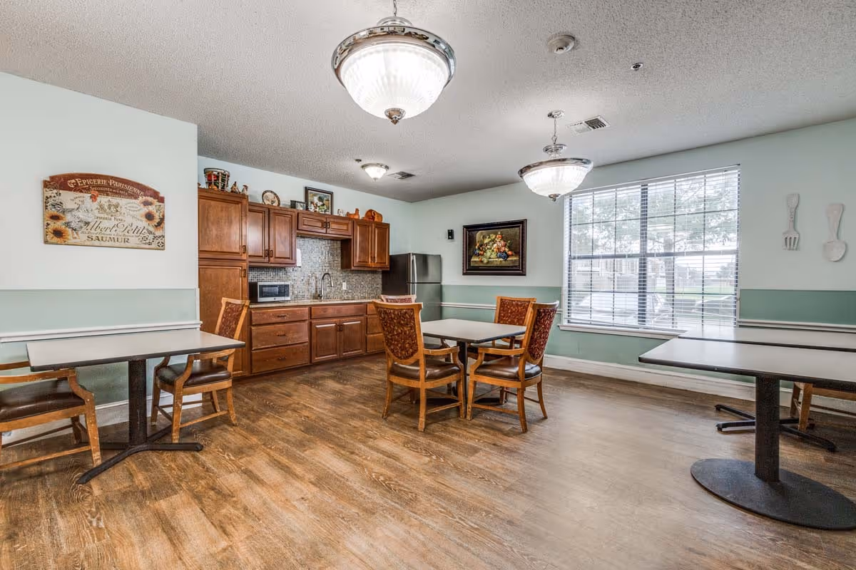 A senior living facility dining area with wooden floors, several tables and chairs, a kitchenette with wooden cabinets, a microwave, and a refrigerator. The room has two large hanging light fixtures, a large window with blinds, and decorative wall art including a framed painting and kitchen utensil decorations.