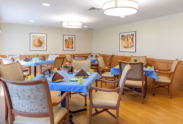 A dining room with several tables covered in blue tablecloths, each set with yellow plates, cups, and brown folded napkins. The room has wooden chairs with patterned cushions, light-colored walls with framed artwork of pears, and a wooden floor. Ceiling lights provide bright illumination.