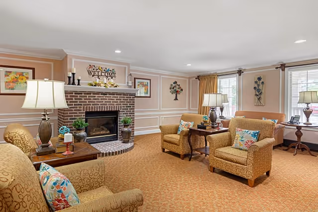 A cozy living room area with beige patterned armchairs and a sofa, colorful floral cushions, wooden side tables with lamps, and a brick fireplace with decorative items and a welcome sign above it. The room has beige walls with white trim, carpeted floor, and windows with curtains allowing natural light.