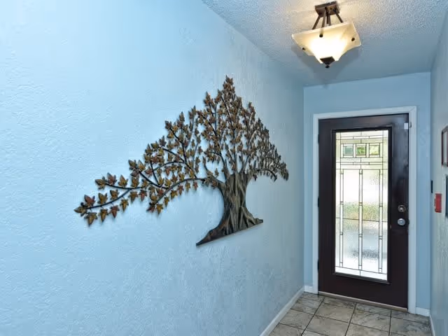 A narrow hallway with light blue walls and tiled floor leading to a door with decorative frosted glass panels. A metal wall art piece shaped like a tree with leaves is mounted on the left wall. A ceiling light fixture is visible above.