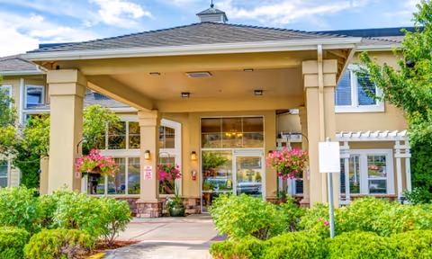 Entrance of a senior living facility with a covered drop-off area supported by large columns, surrounded by green shrubs and hanging flower baskets. The building has large windows and a beige exterior with a peaked roof.
