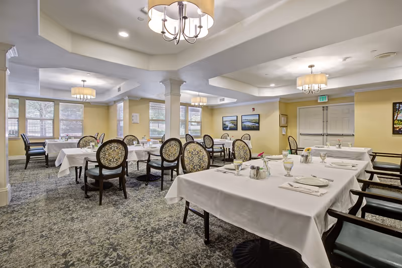 A bright and spacious dining room with multiple tables covered in white tablecloths, each set with plates, glasses, napkins, and silverware. The room features patterned upholstered chairs, large windows letting in natural light, beige walls, carpeted floors, and ceiling light fixtures.