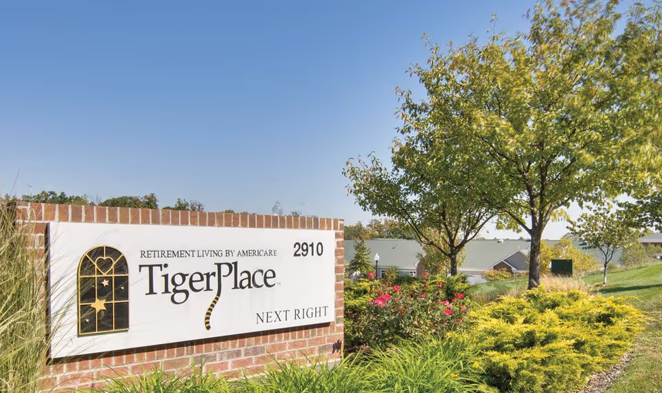 Entrance sign reading 'TigerPlace' on a brick wall with landscaped shrubs, trees, and building roofs in the background.