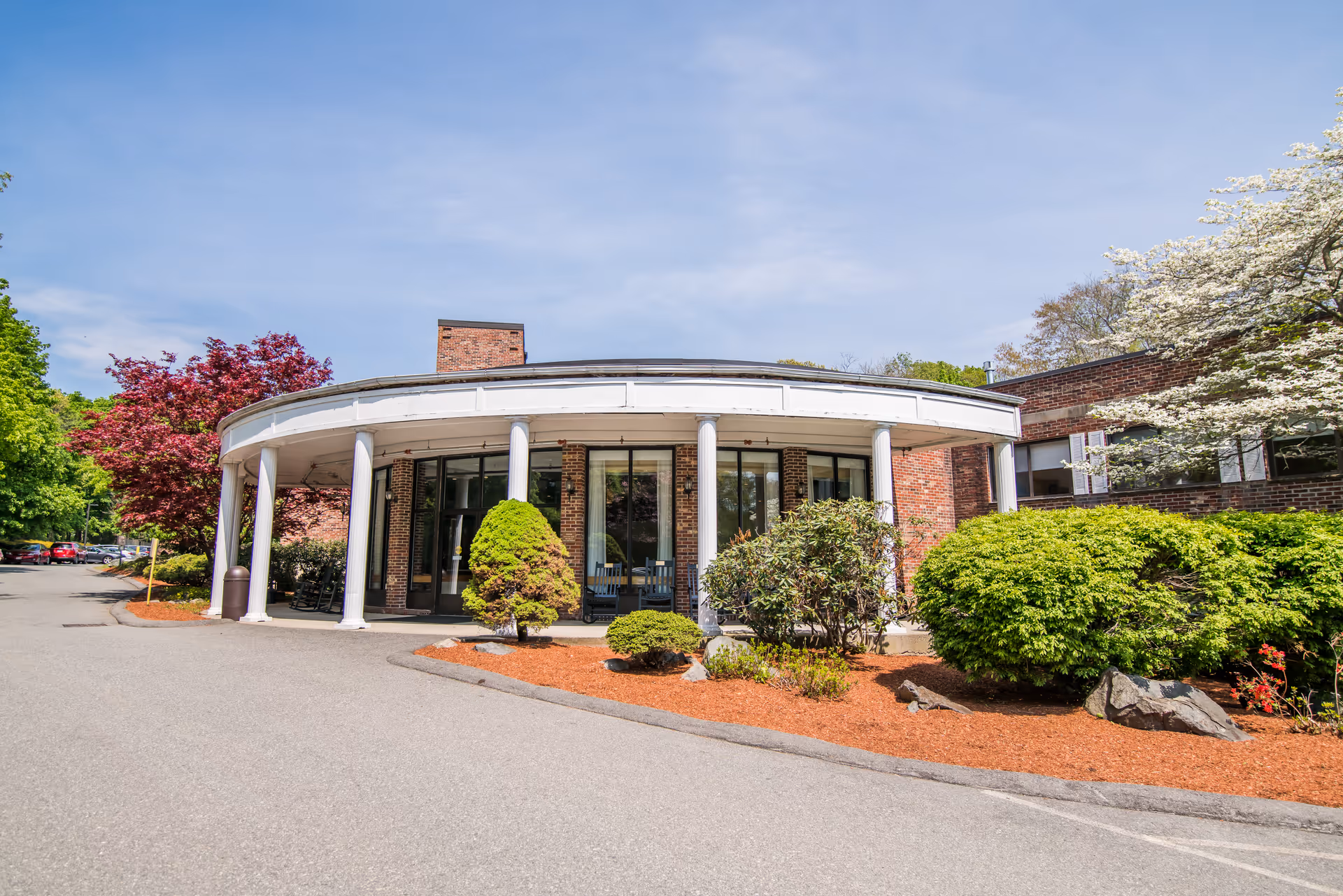 Front exterior view of a brick building with a curved white portico supported by columns, surrounded by landscaped bushes and trees under a clear blue sky.