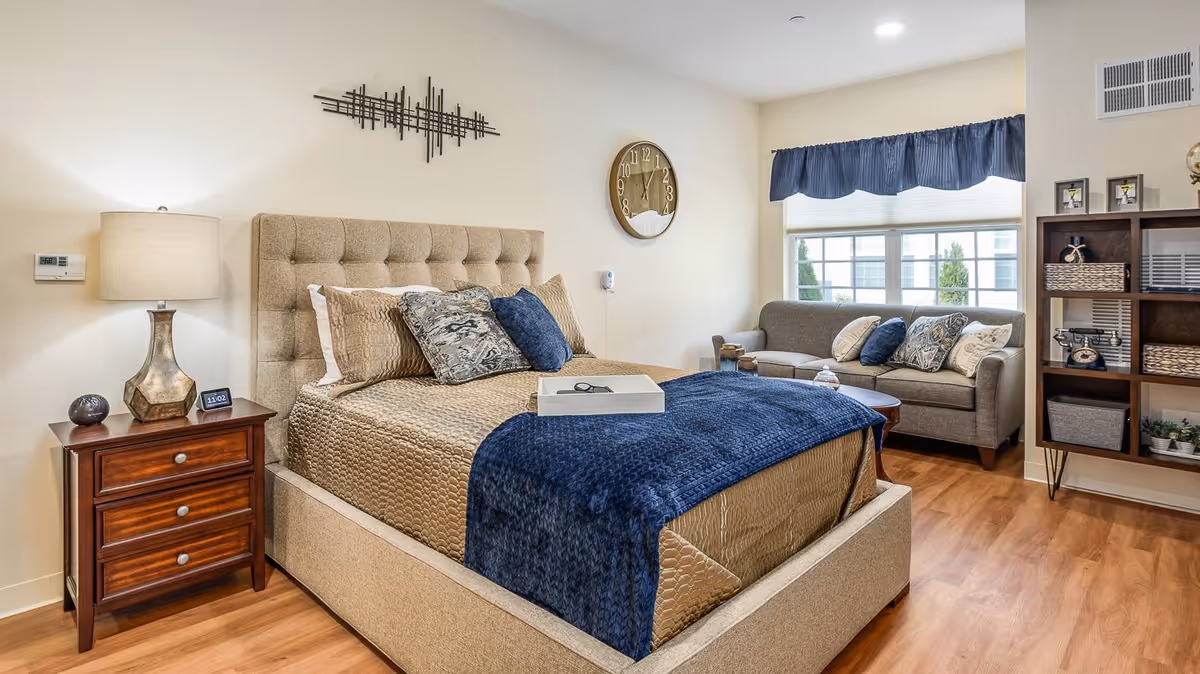 A cozy bedroom featuring a beige upholstered bed with multiple pillows and a blue throw blanket. Next to the bed is a wooden nightstand with a lamp, a small clock, and decorative items. A gray sofa with decorative pillows is placed near a window with blue valance curtains. A wooden shelving unit with baskets and decor is on the right side of the room. The floor is wooden, and the walls are painted light beige.
