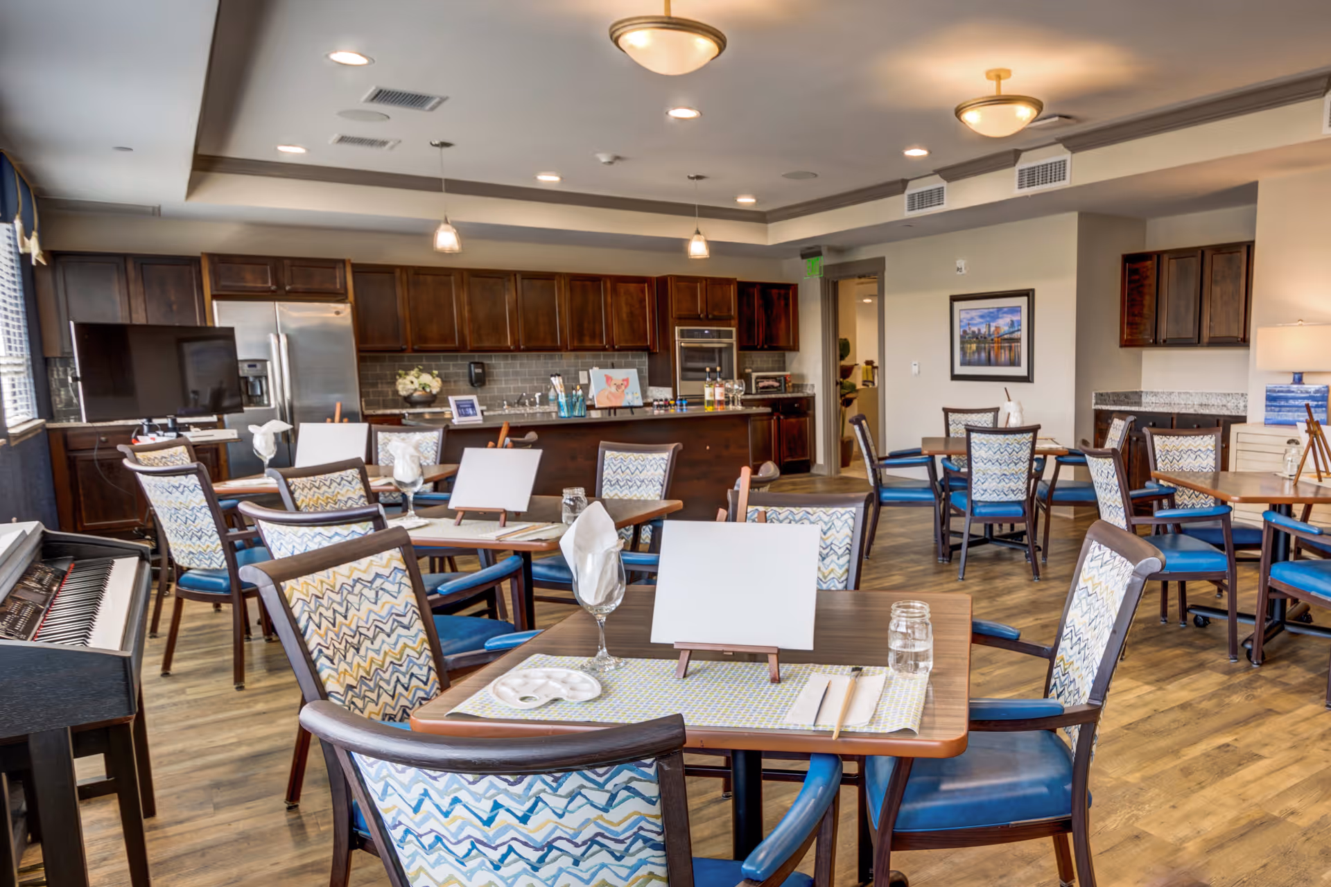 A bright dining room with multiple wooden tables and chairs featuring blue and patterned upholstery. Each table is set with placemats, napkins, glasses, and small easels with blank cards. The room has wooden flooring, a piano on the left side, and a kitchen area with dark wooden cabinets, a stainless steel refrigerator, and a countertop in the background. Ceiling lights illuminate the space, and a framed picture hangs on the far wall.