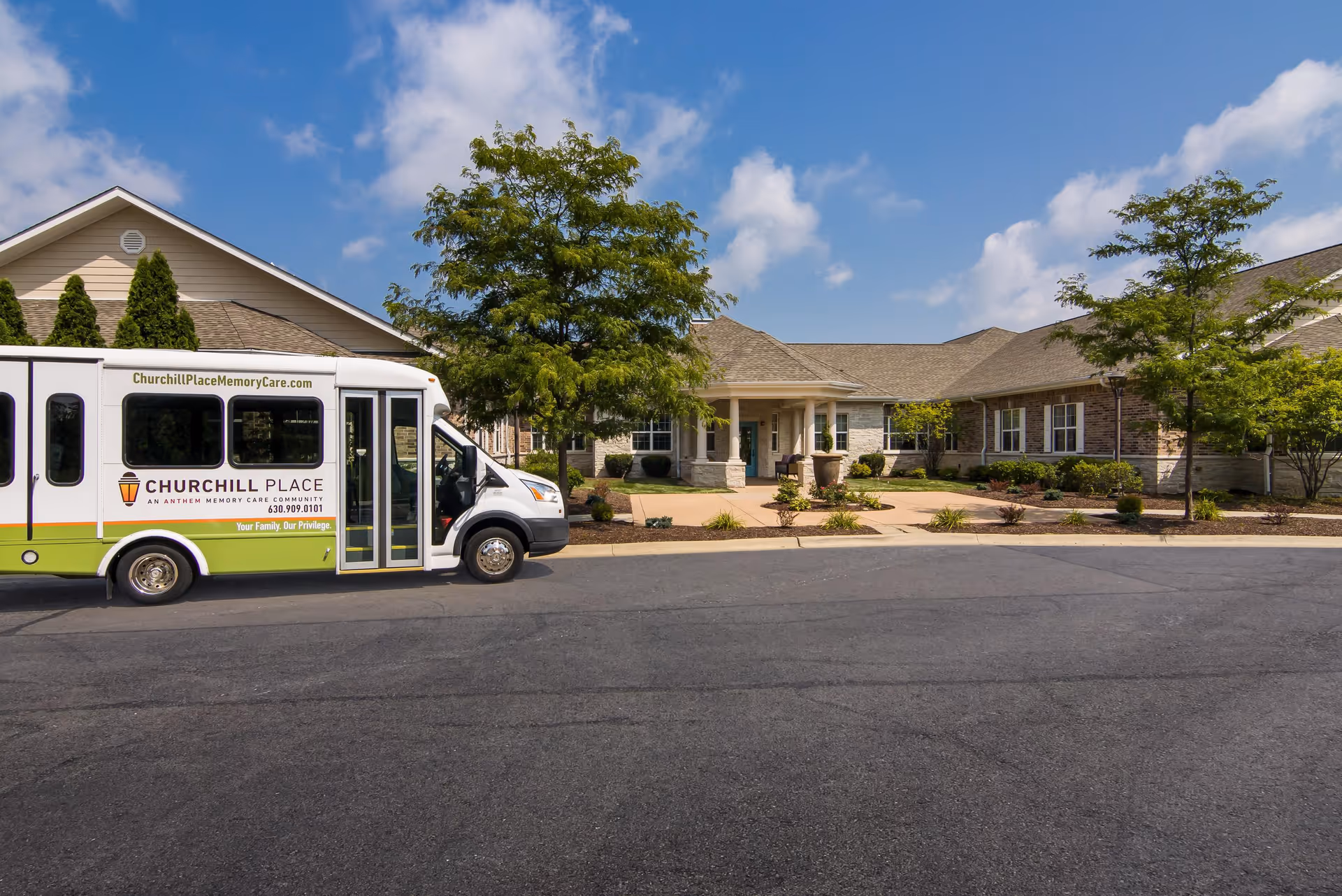 Exterior view of Churchill Place Memory Care facility with a shuttle bus parked in front. The building has a beige and brick facade with a covered entrance, surrounded by landscaped greenery and trees under a partly cloudy sky.