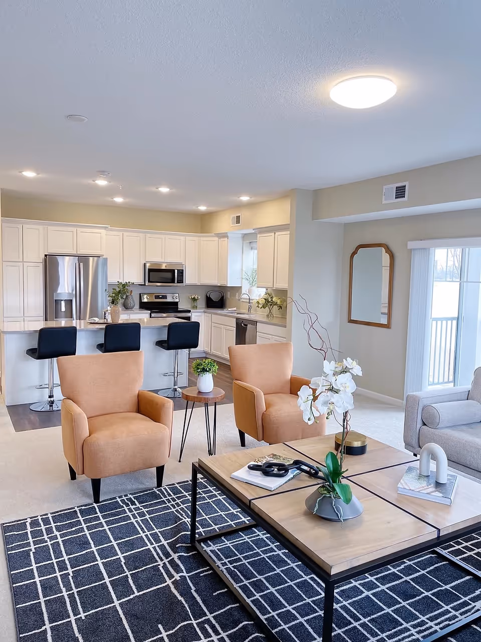 A modern open-concept living area featuring a kitchen with white cabinets, stainless steel appliances, and a breakfast bar with three black bar stools. In the foreground, there are two tan armchairs, a wooden coffee table with decorative items including a white orchid plant, and a black and white patterned rug. A large window with vertical blinds and a wall mirror are visible in the background.