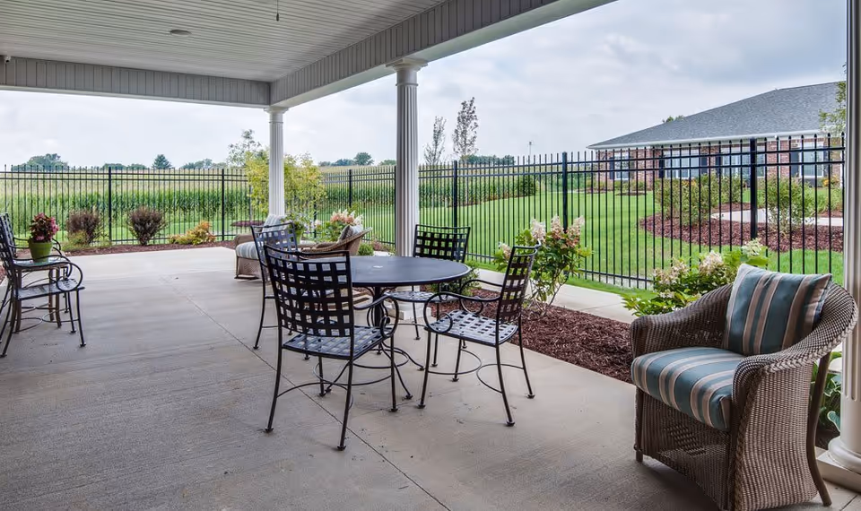 Covered outdoor patio area with metal table and chairs, wicker armchair with striped cushion, potted plants, and a view of a fenced green lawn and a building in the background under a cloudy sky.