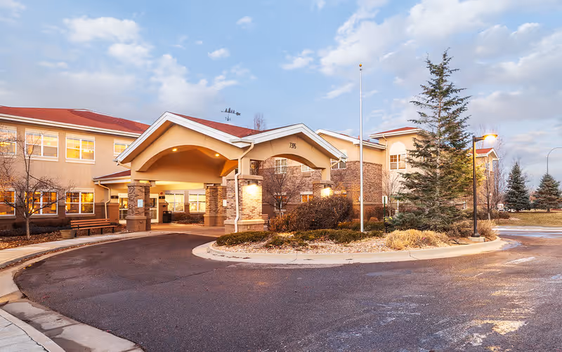 Exterior view of Lakewood Post Acute and Rehabilitation facility showing the main entrance with a covered driveway, surrounded by trees and landscaping under a partly cloudy sky.
