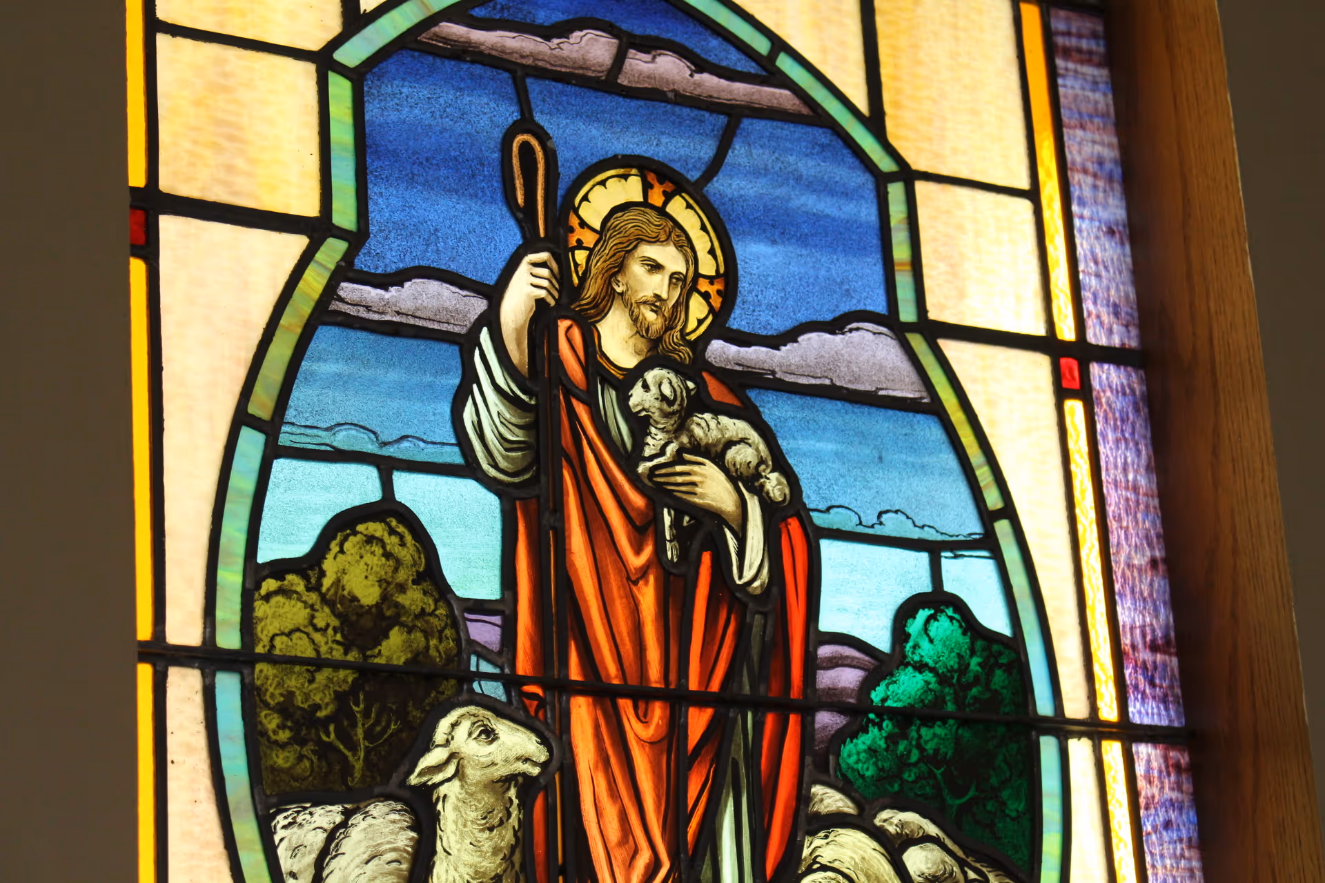Stained glass window showing Jesus as the Good Shepherd holding a lamb with other sheep and a landscape background.