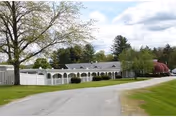 Single-story white nursing facility with an arched covered walkway, white fence, driveway, and surrounding trees and lawn under a partly cloudy sky.