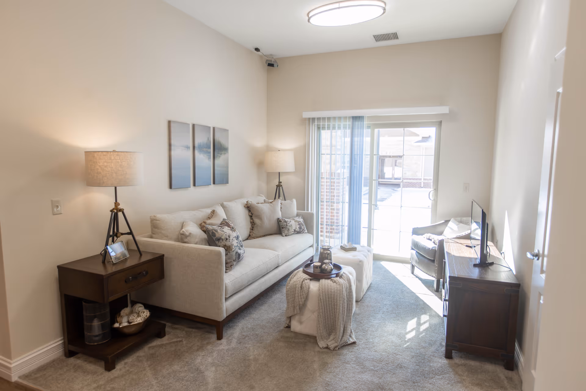 A bright living room with a beige sofa adorned with patterned cushions, a wooden side table with a lamp and photo frame, a small ottoman with a tray and blanket, a gray armchair, a wooden TV stand with a flat-screen TV, and a sliding glass door with vertical blinds letting in natural light.