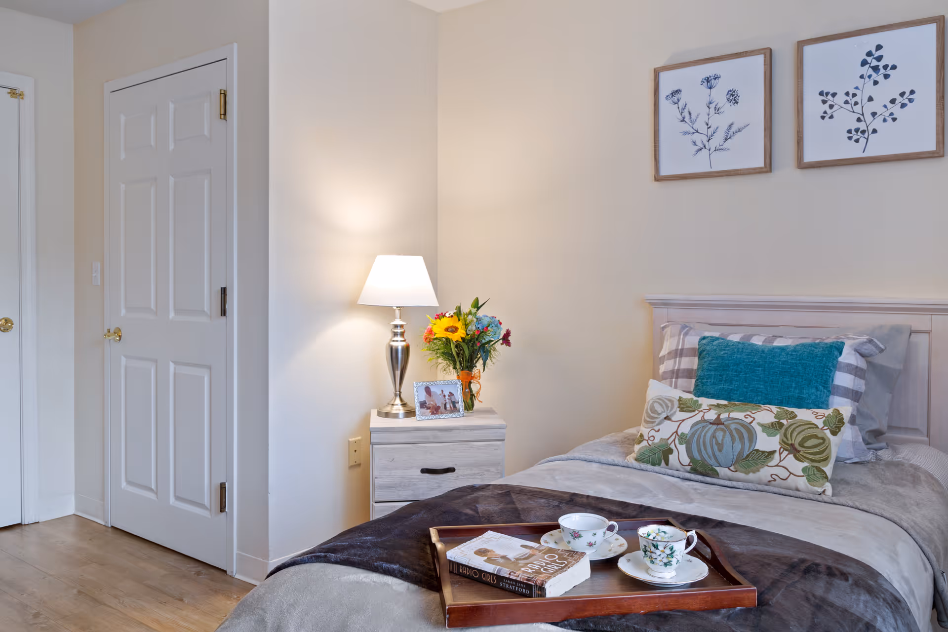 A cozy bedroom with a single bed featuring multiple pillows and a blanket. A wooden tray with two teacups and a book rests on the bed. Next to the bed is a nightstand with a silver lamp, a small bouquet of flowers, and a framed photo. Two framed botanical prints hang on the wall above the bed. The room has light-colored walls and wooden flooring, with two closed white doors visible on the left side.
