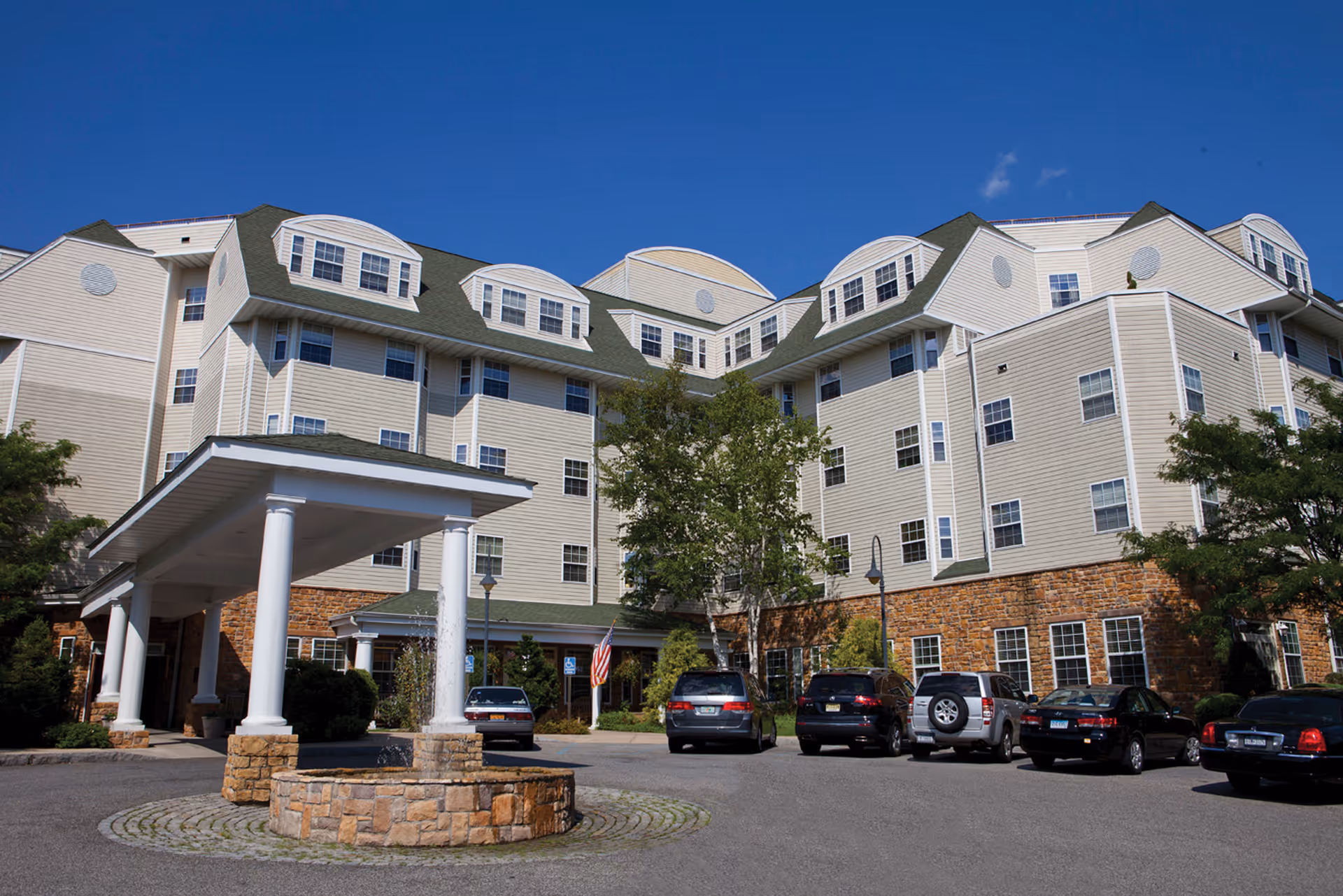 Front entrance of a multi-story senior living building with a covered porte-cochere, circular stone fountain, and parked cars.