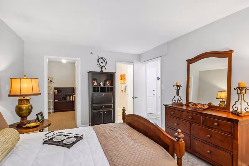 A cozy bedroom with a wooden bed frame, a brown quilt, and a white bedspread. There is a bedside table with a lamp and a small picture frame. A wooden dresser with a large mirror and decorative candle holders is on the right side. Two open doors reveal a walk-in closet with shelves and a bathroom with a white door. The walls are painted light gray and the room is well-lit with natural and artificial light.