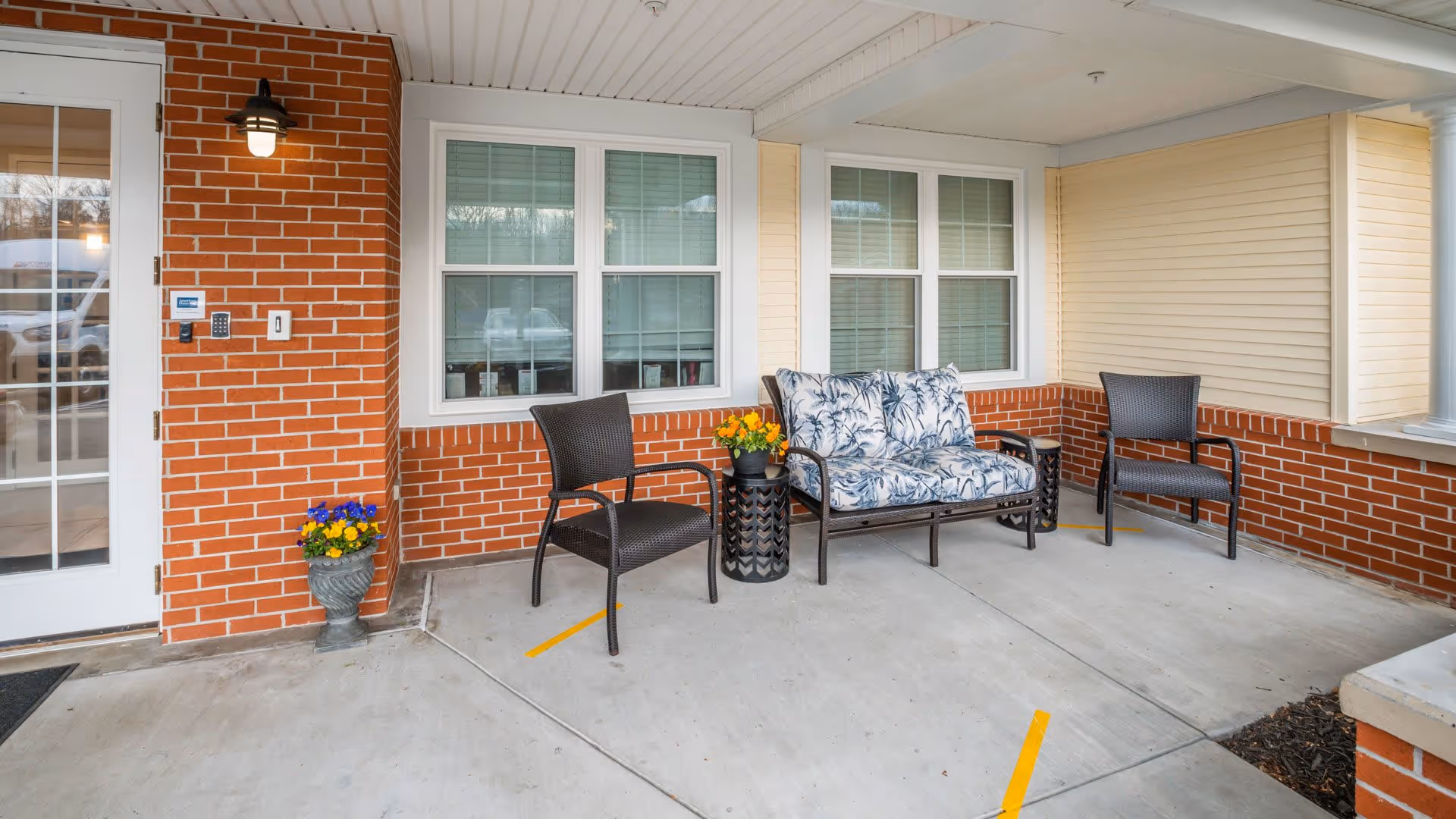 Covered outdoor seating area with two black chairs and a cushioned loveseat with a floral pattern, placed on a concrete floor next to a brick and beige siding wall with two windows. There are two small flower pots with colorful flowers near the seating.