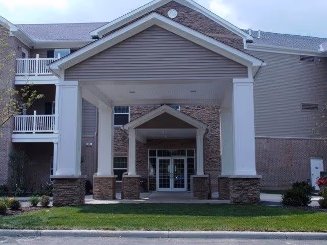 Front exterior view of a multi-story building with a covered entrance supported by large white columns and stone bases. The building has beige siding and brick accents, with balconies visible on the left side.