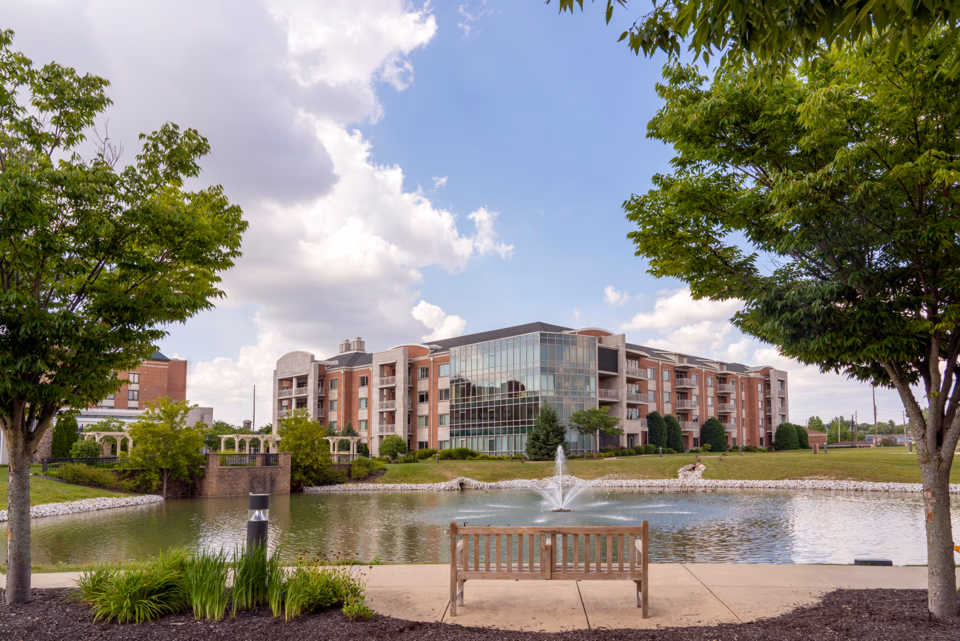 View of a senior living facility named Marquette with a large pond and fountain in the foreground, a wooden bench on a paved path, and green trees framing the scene under a partly cloudy sky.