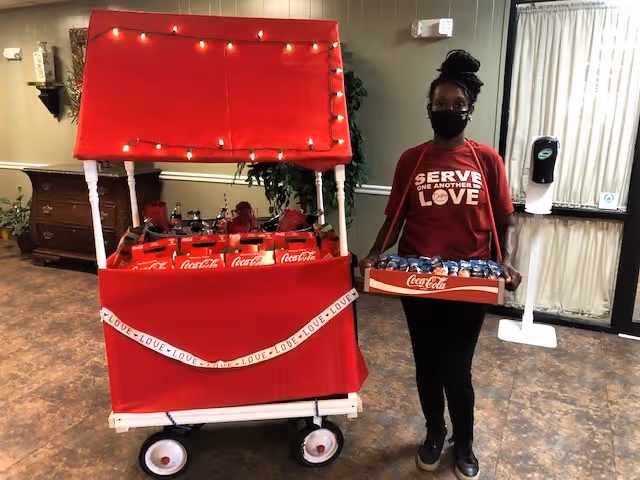 A person wearing a red shirt with the words 'SERVE ONE ANOTHER LOVE' and a black face mask stands indoors holding a tray of Coca-Cola cans. Next to them is a red cart decorated with string lights and a ribbon that says 'LOVE' repeatedly, filled with Coca-Cola bottles. The room has a brown floor, a wooden dresser, plants, and a hand sanitizer dispenser near a window with curtains.