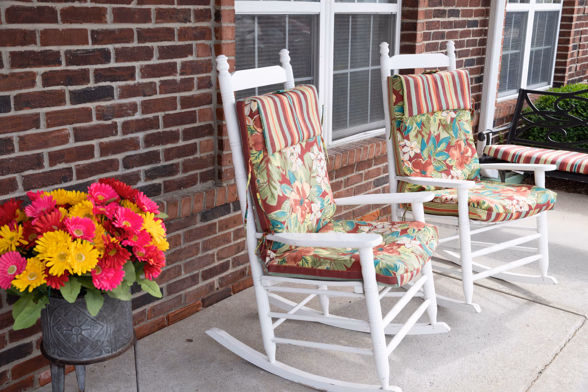 Two white wooden rocking chairs with floral and striped cushions placed on a concrete porch in front of a brick wall with windows. To the left, there is a metal planter filled with bright pink and yellow flowers. A black metal bench with a striped cushion is partially visible on the right side.