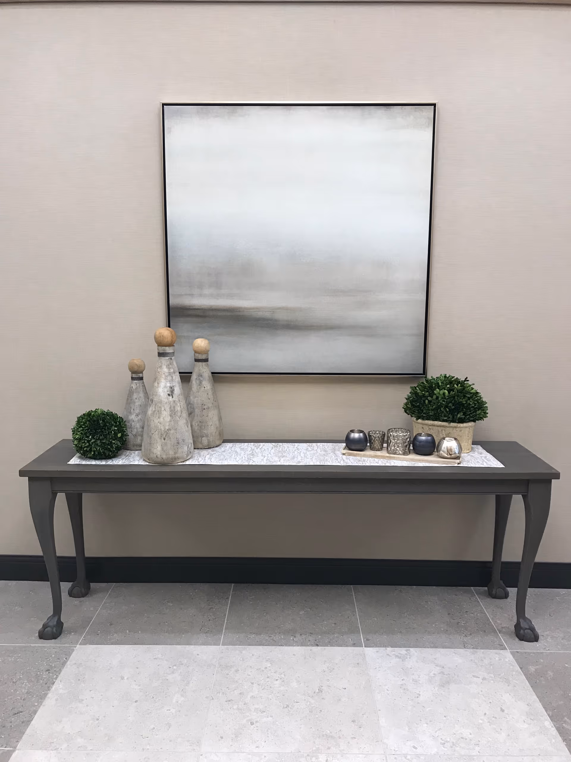 A decorative console table with claw feet against a beige wall. On the table are three rustic wooden bottle-shaped sculptures with round tops, two small green potted plants, and a tray holding several small candle holders. Above the table hangs a large abstract painting in shades of gray and white.