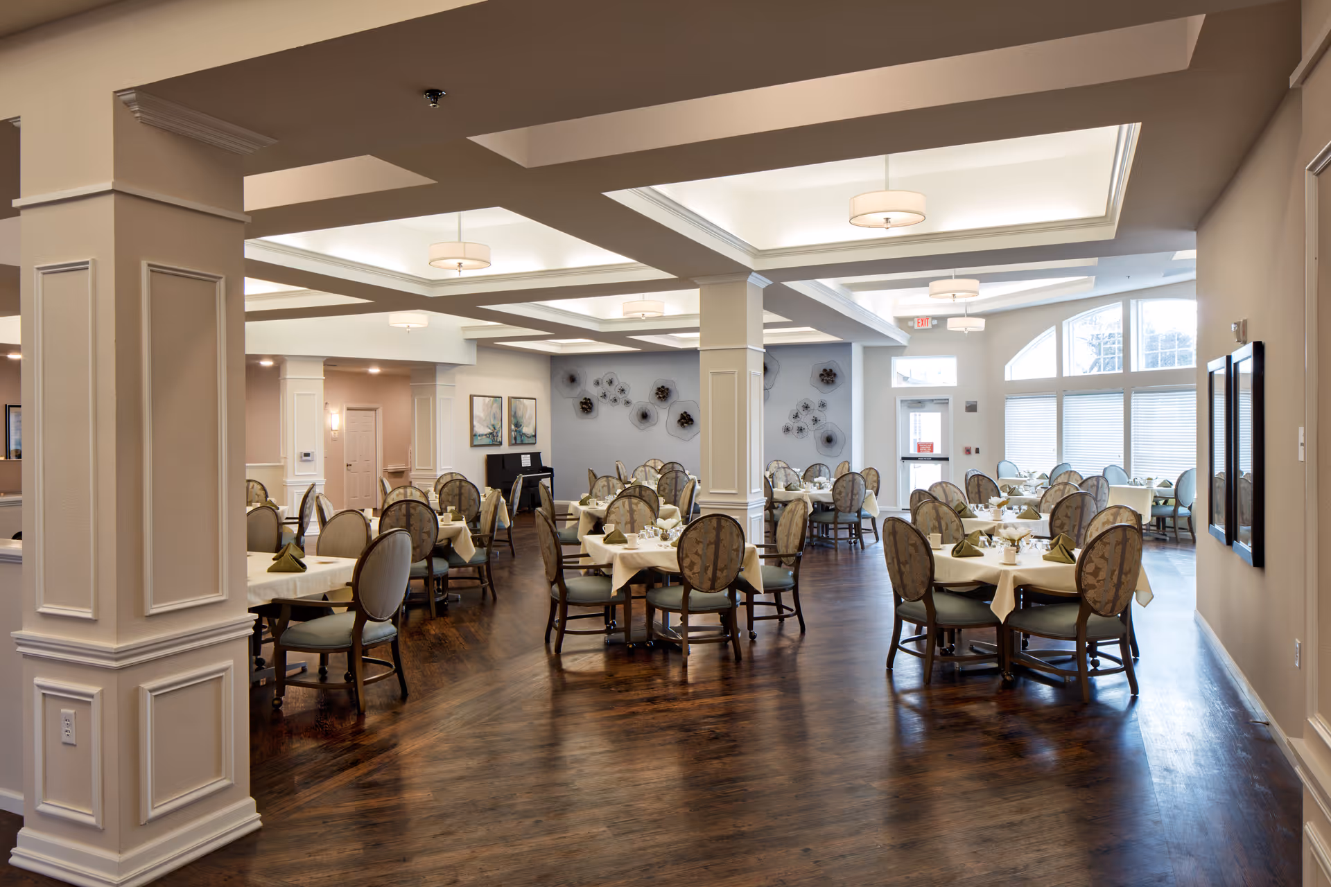 A spacious dining room with multiple round tables covered with white tablecloths and set with napkins and tableware. The room features elegant upholstered chairs, wooden flooring, decorative wall art, and large windows allowing natural light to fill the space.