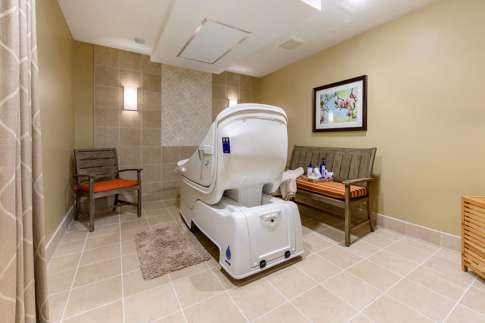 A serene room with beige tiled floor and walls, featuring a white hydrotherapy bath in the center. There are two wooden benches with orange cushions on either side of the bath, one holding towels and bath products. A framed floral picture hangs on the wall above one bench, and soft lighting fixtures illuminate the space.