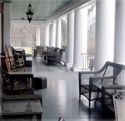 Wide covered porch with wicker chairs and pillars, overlooking a grassy lawn.