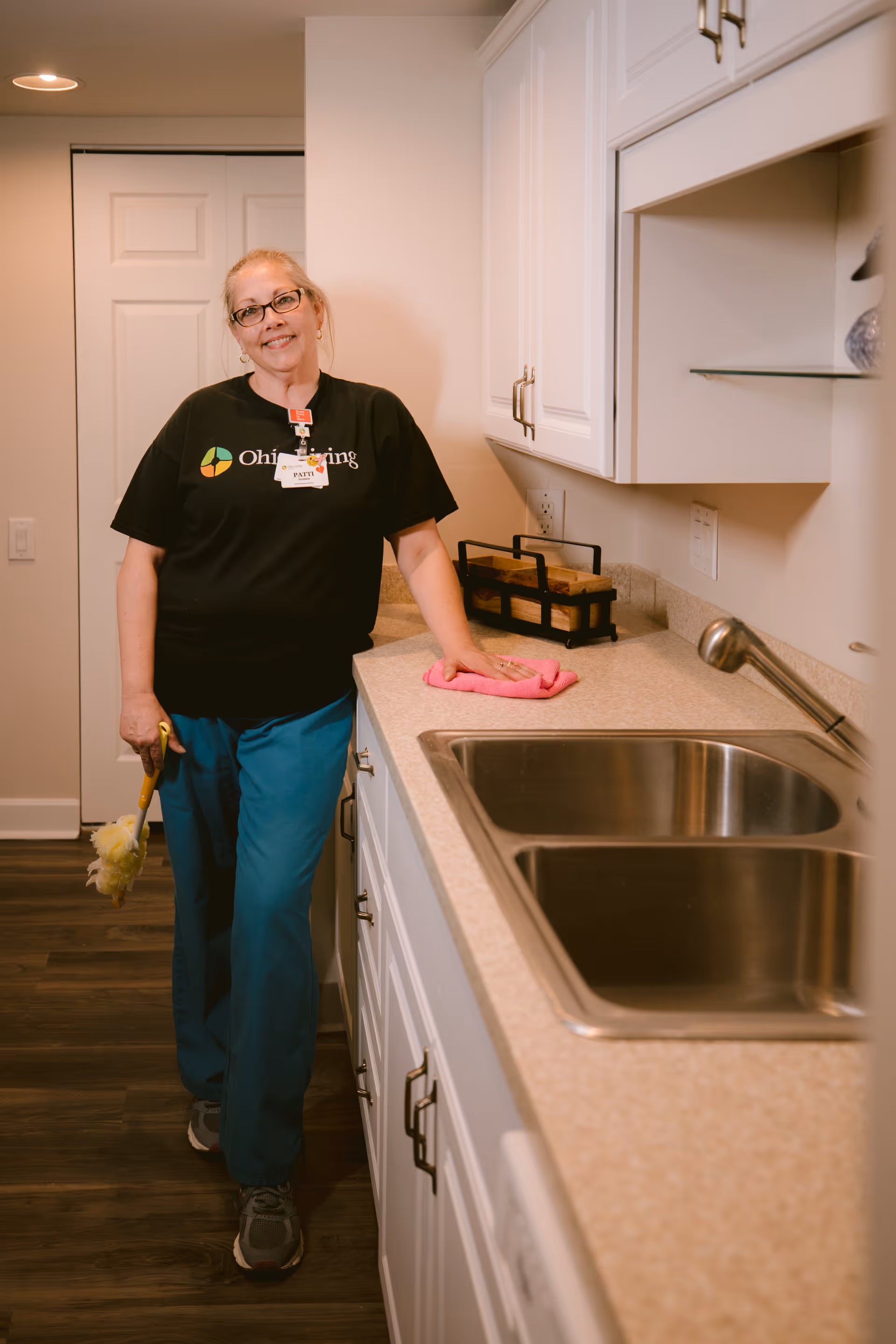 A woman wearing glasses and a black Ohio Living shirt stands in a kitchen next to a countertop with a double sink. She is holding a yellow cleaning brush in one hand and resting the other hand on the counter with a pink cleaning cloth. The kitchen has white cabinets and a wooden floor.