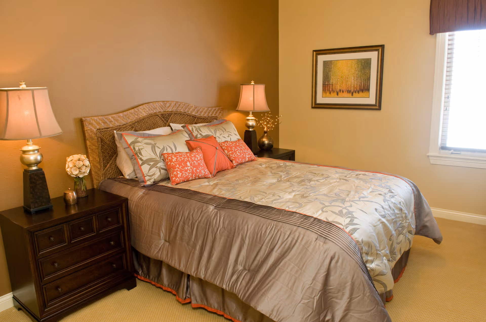 A neatly made bed with a decorative headboard and multiple pillows in a warmly lit bedroom. There are two bedside tables, each with a lamp and decorative items. A framed picture hangs on the wall next to a window with blinds partially open.