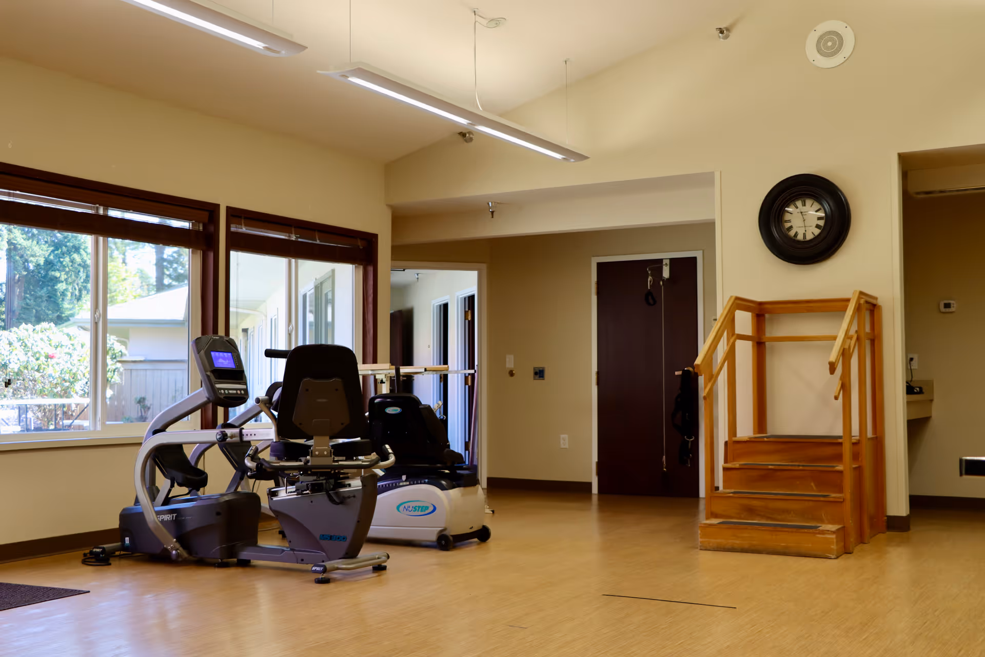 A bright interior fitness/activity room with exercise machines, a small wooden practice staircase, a wall clock, and large windows.