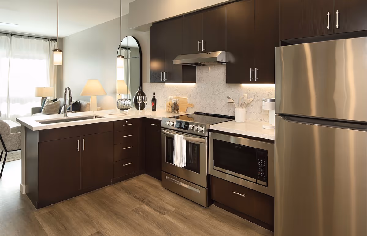 Modern kitchen with dark wood cabinets, stainless steel refrigerator, oven, and microwave. The kitchen has a white marble countertop with a sink and faucet, decorative jars, a bottle of wine, and kitchen utensils. A mirror and a lamp are visible in the adjacent living area.