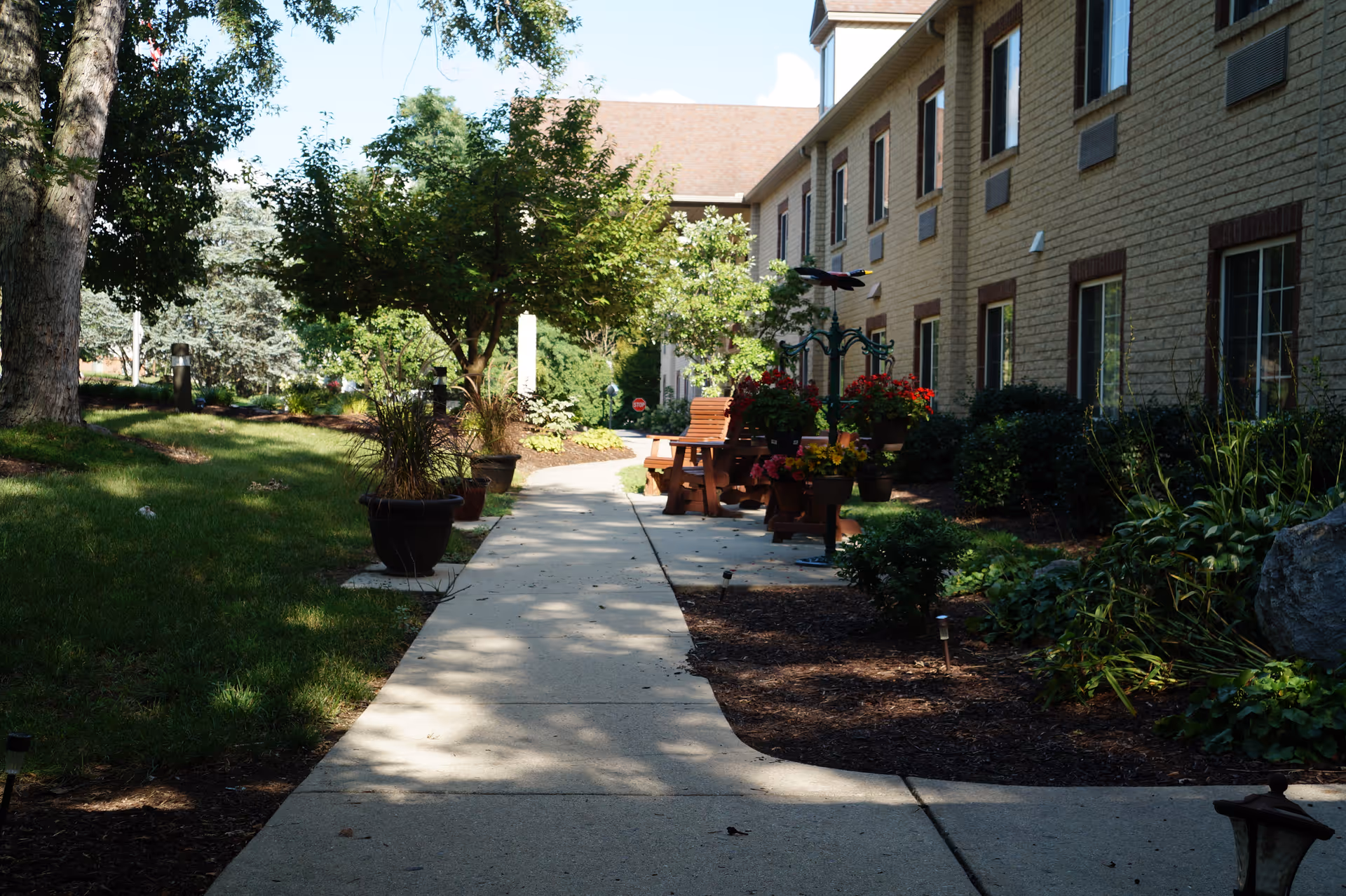 A paved walkway lined with potted plants and flowers leading alongside a two-story brick building. There are benches and a decorative metal arch with hanging flower baskets along the path. Trees and shrubs provide shade and greenery on both sides of the walkway.
