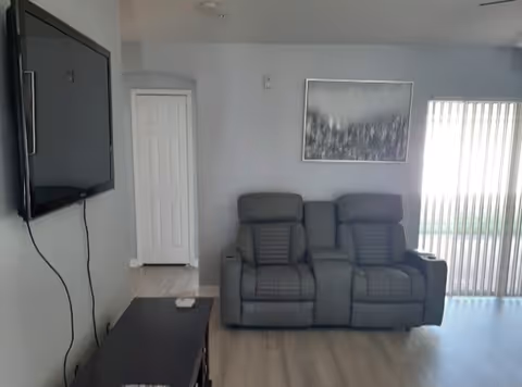 A living room area with two gray recliner chairs positioned side by side against a light gray wall. Above the chairs hangs a framed abstract painting. To the left, a flat-screen TV is mounted on the wall with visible cords hanging down, and below it is a dark wooden cabinet. On the right side, vertical blinds cover a sliding glass door allowing natural light into the room. The floor is light-colored wood.