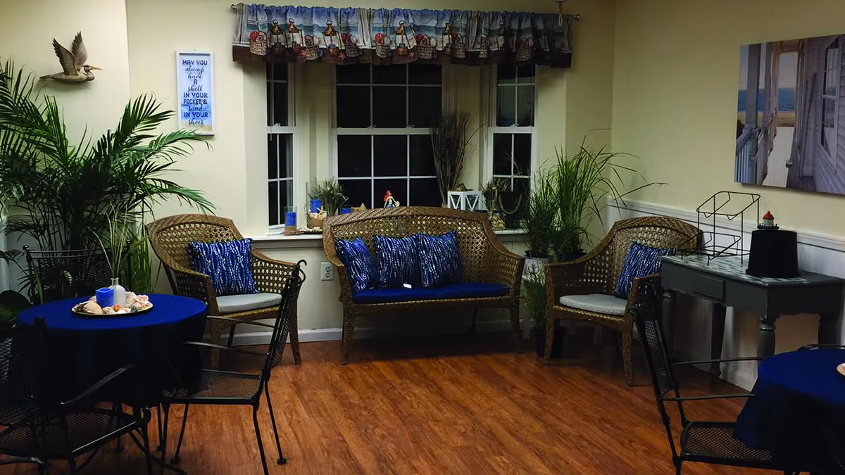 A cozy communal room with wicker seating and blue-covered round tables, potted plants, and a large window.