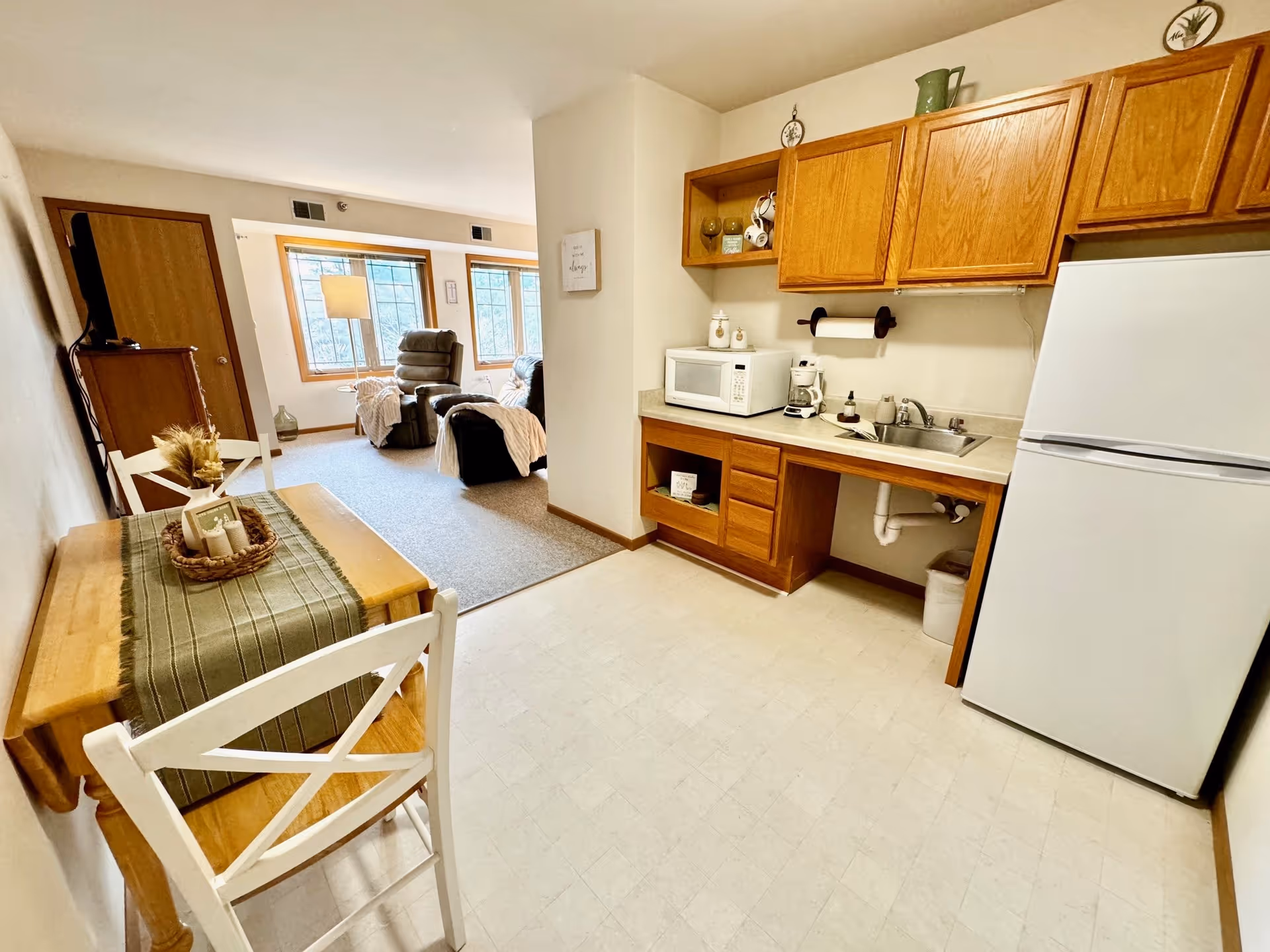 A small kitchen area with wooden cabinets, a white refrigerator, microwave, coffee maker, and a sink. Adjacent to the kitchen is a dining table with two white chairs and a green table runner with decorative items. In the background, there is a living room area with carpeted flooring, two recliner chairs, a floor lamp, and large windows letting in natural light.